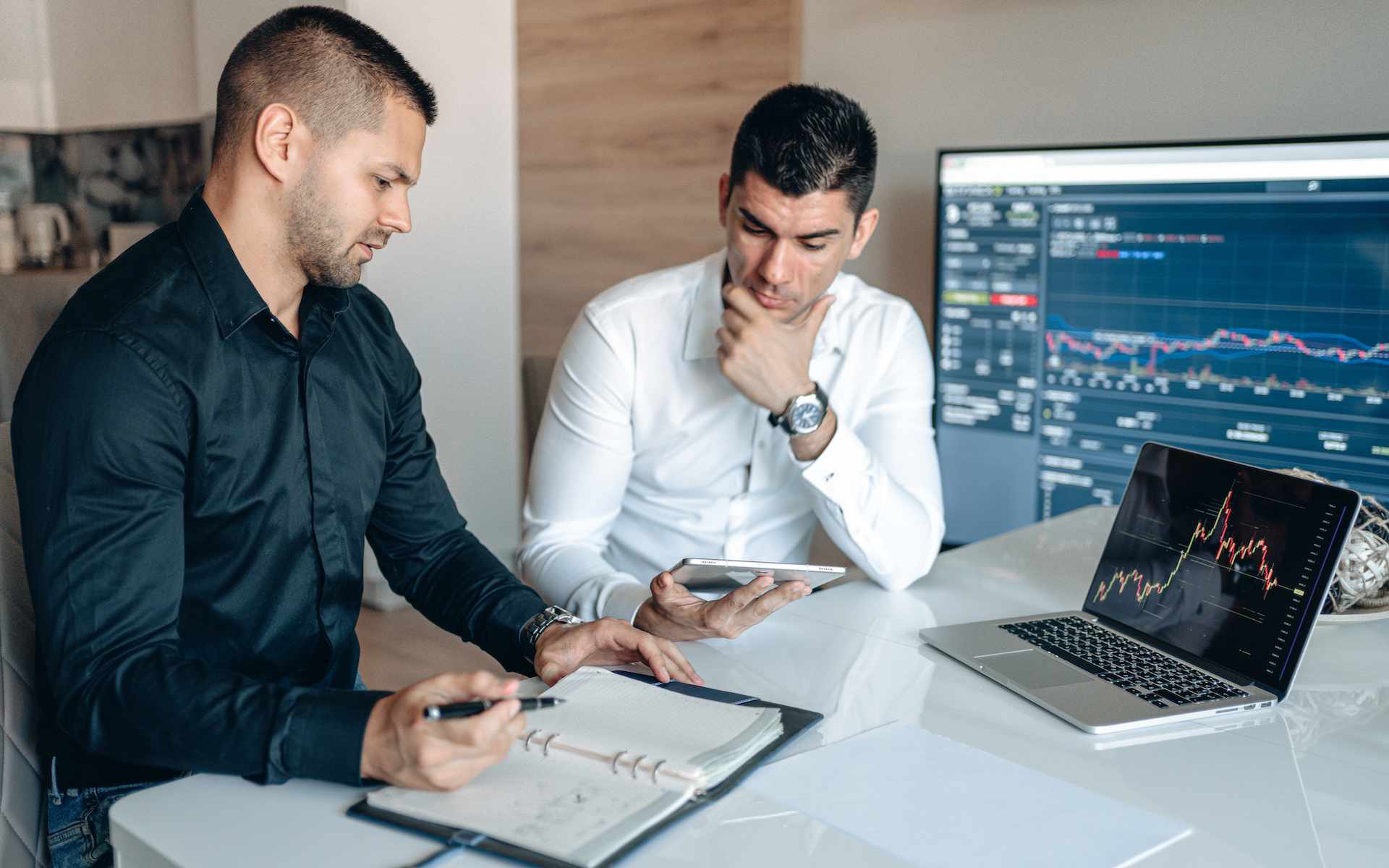 Two men are sitting at a table looking at an iPad. On the table is a laptop on which a diagram can be seen.